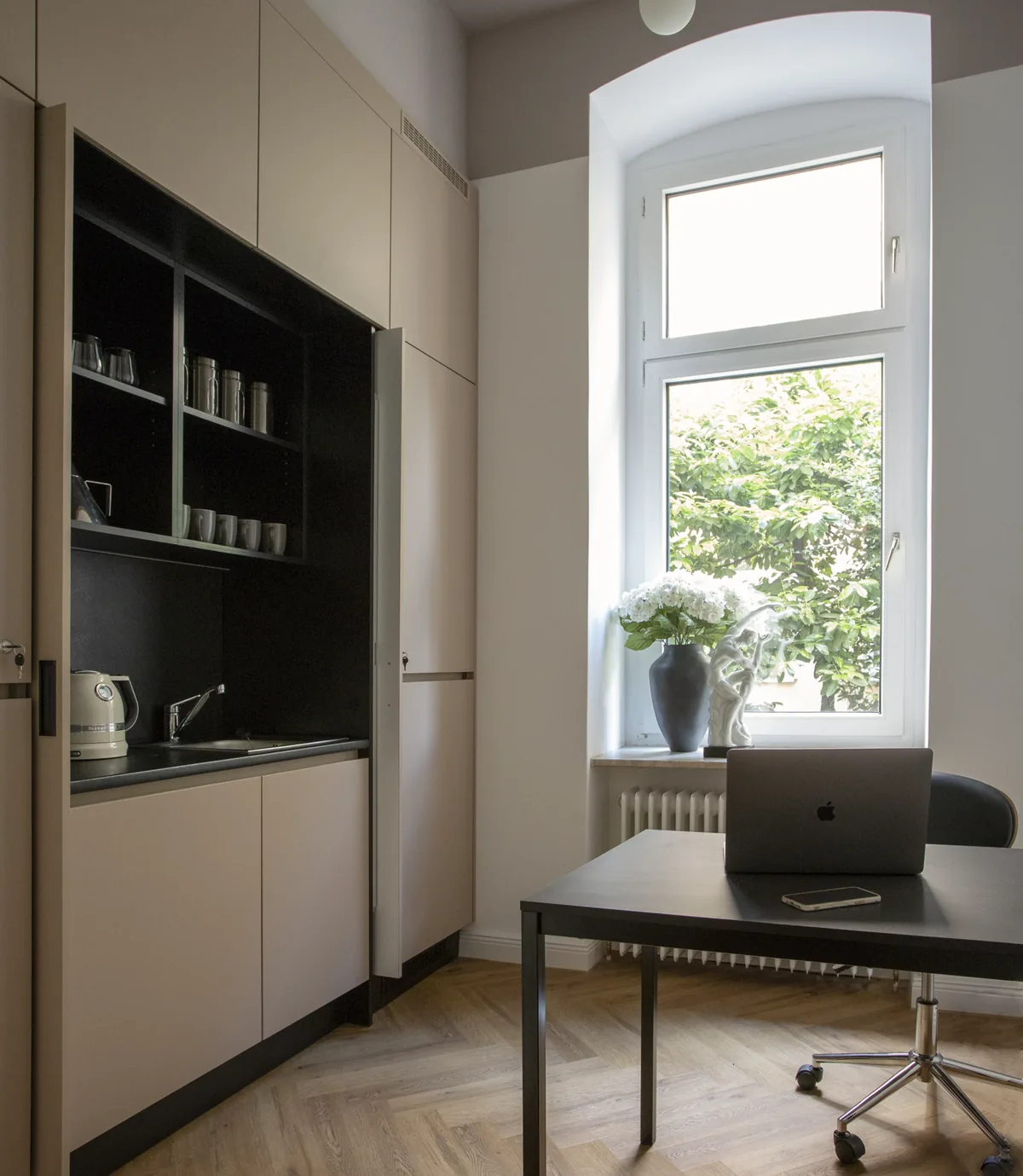 Zimmer mit eingebauter Küchennische, einem schwarzen Schreibtisch mit Laptop und einem Fenster mit Blumen auf der Fensterbank.