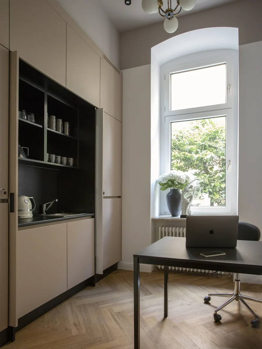 Zimmer mit eingebauter Küchennische, einem schwarzen Schreibtisch mit Laptop und einem Fenster mit Blumen auf der Fensterbank.