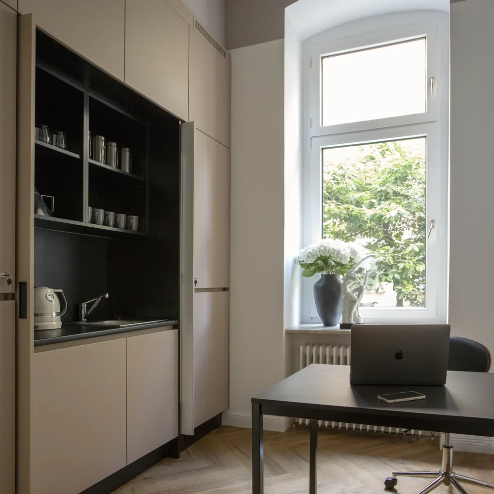 Zimmer mit eingebauter Küchennische, einem schwarzen Schreibtisch mit Laptop und einem Fenster mit Blumen auf der Fensterbank.