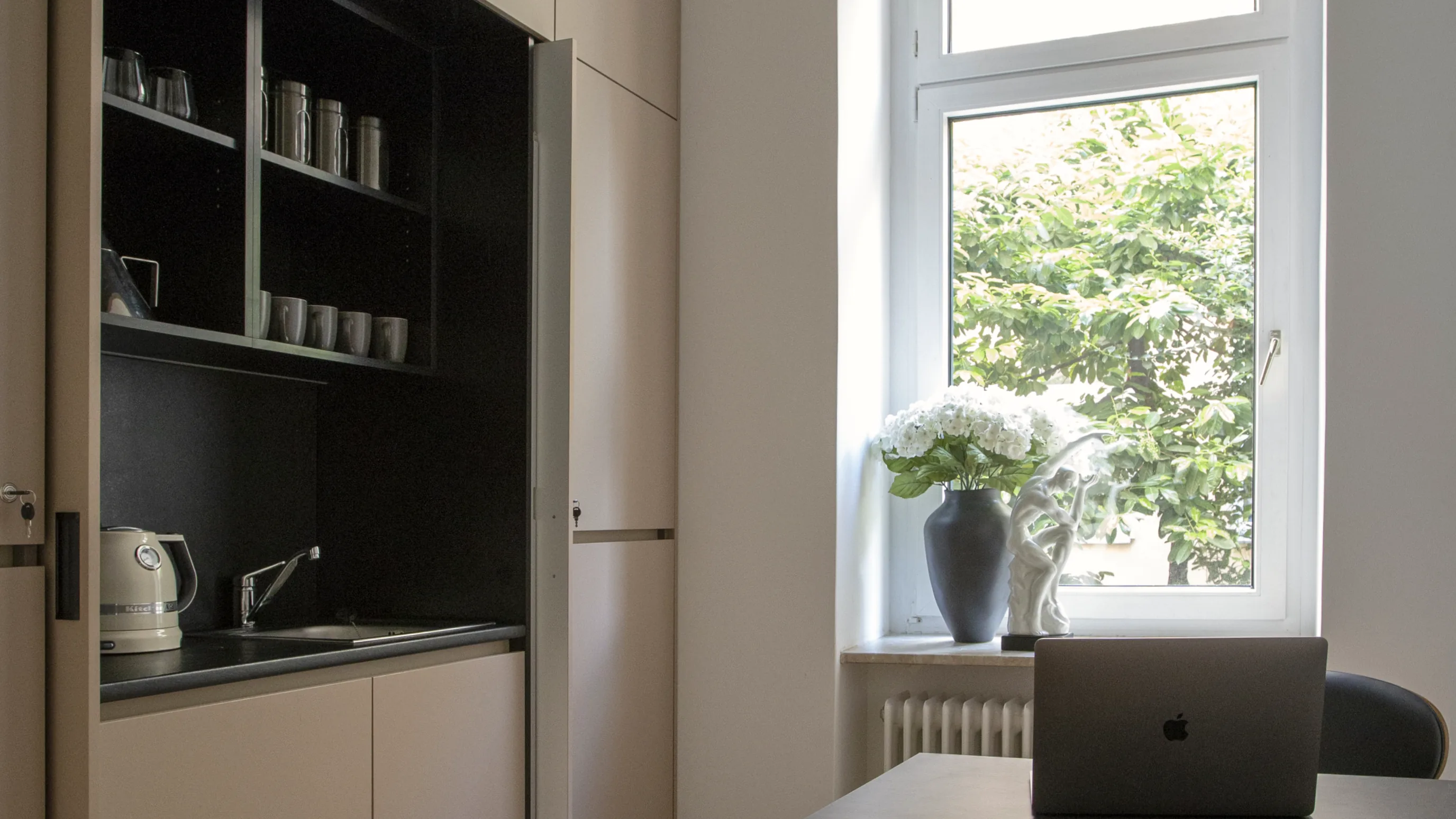Zimmer mit eingebauter Küchennische, einem schwarzen Schreibtisch mit Laptop und einem Fenster mit Blumen auf der Fensterbank.