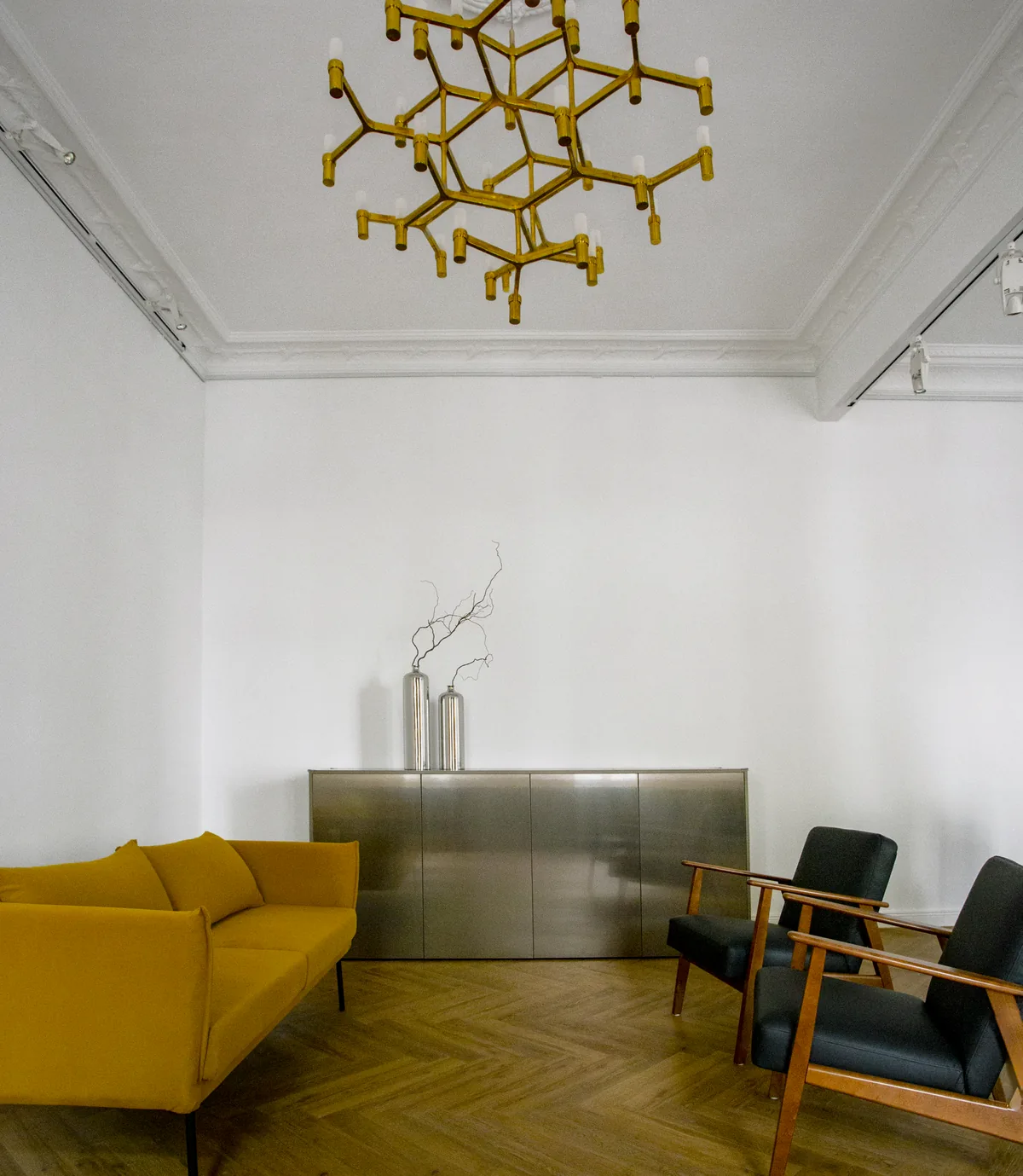 Ein Raum mit einem Messing-Kronleuchter an der Decke, einem gelben Sofa, einem Metallschrank und zwei Sesseln auf Fischgrätparkett