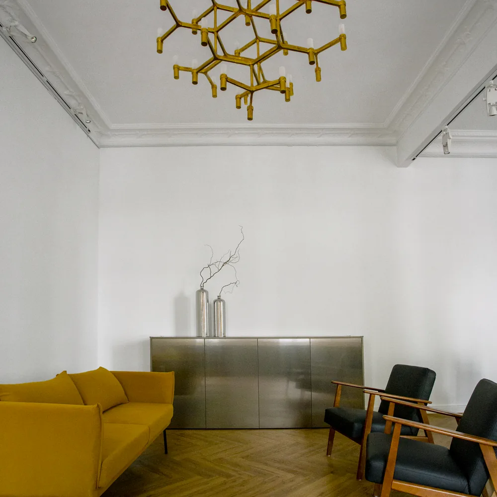 Ein Raum mit einem Messing-Kronleuchter an der Decke, einem gelben Sofa, einem Metallschrank und zwei Sesseln auf Fischgrätparkett