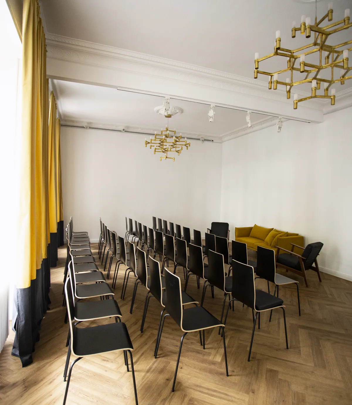 Stuhlreihen aus schwarzen Stühlen im Saal aus der Perspektive des Fensters, zwei Messing-Kronleuchter an der Decke, ein gelbes Sofa in der Ecke