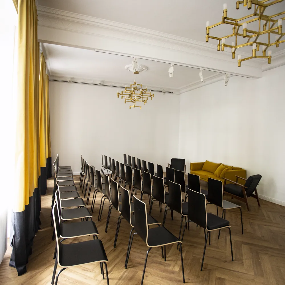 Stuhlreihen aus schwarzen Stühlen im Saal aus der Perspektive des Fensters, zwei Messing-Kronleuchter an der Decke, ein gelbes Sofa in der Ecke