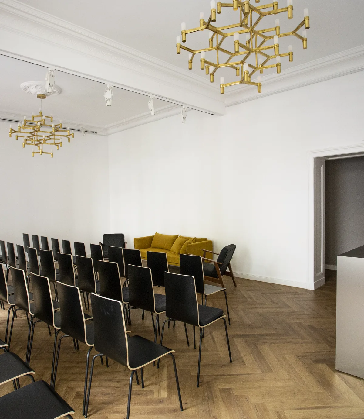 Stuhlreihen aus schwarzen Stühlen im Saal, ein gelbes Sofa im Hintergrund, rechts eine Metalltheke mit Laptop und Vasen