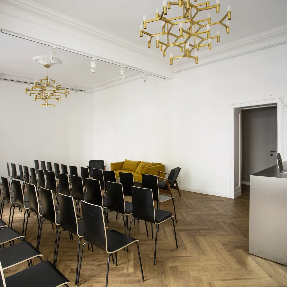 Stuhlreihen aus schwarzen Stühlen im Saal, ein gelbes Sofa im Hintergrund, rechts eine Metalltheke mit Laptop und Vasen