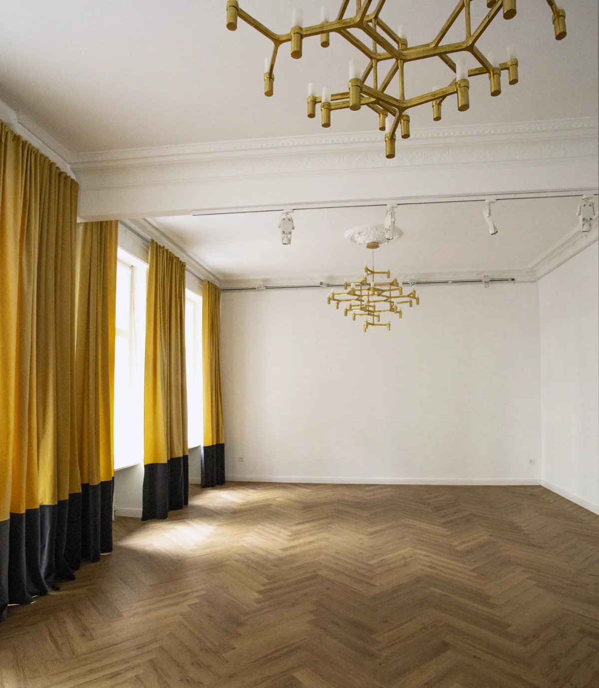 Ein leerer Saal ohne Möbel, Fischgrätboden, Vorhänge oben gelb und unten schwarz, zwei Messing-Kronleuchter an der Decke