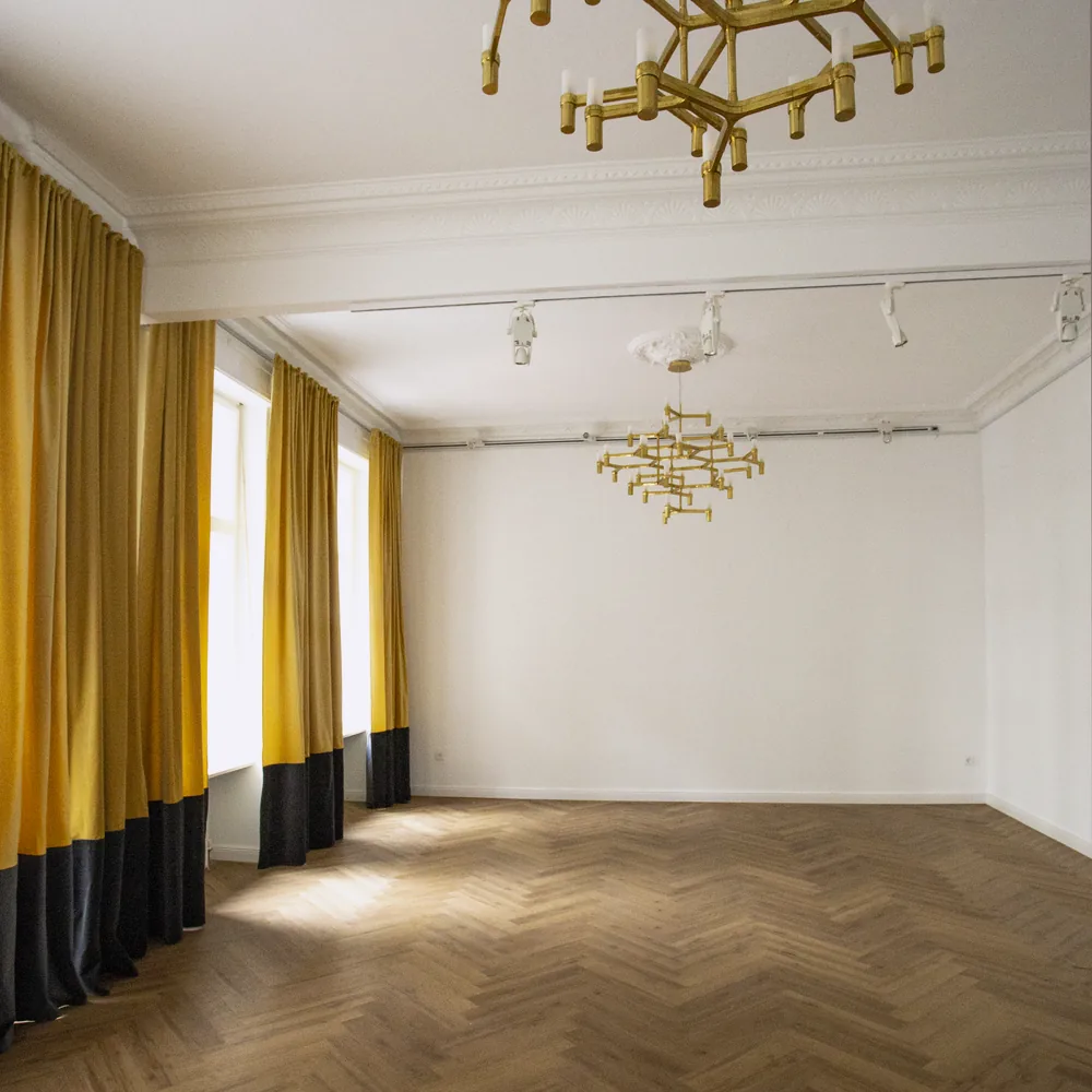 Ein leerer Saal ohne Möbel, Fischgrätboden, Vorhänge oben gelb und unten schwarz, zwei Messing-Kronleuchter an der Decke