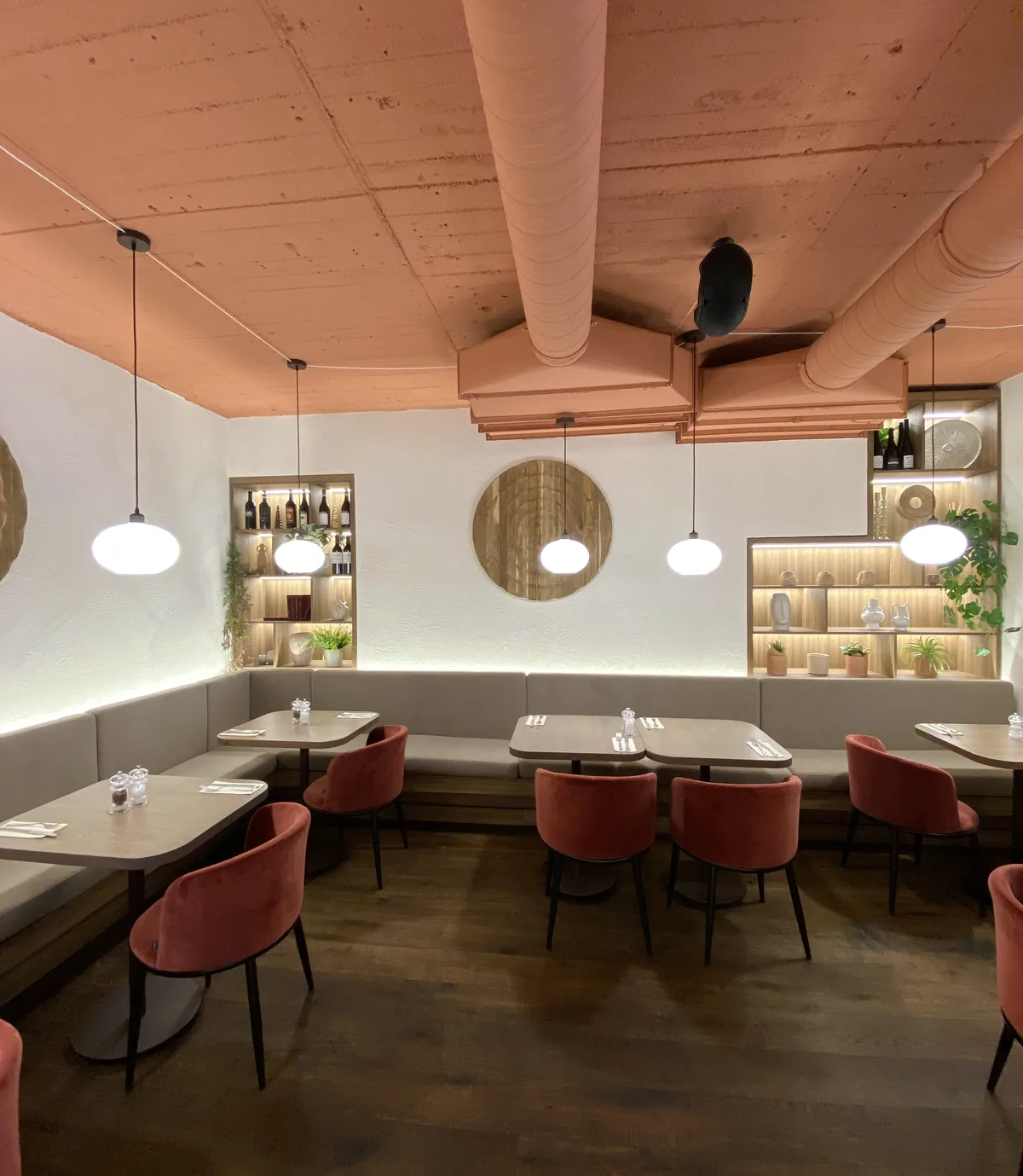 Café-Saal mit einer Tischreihe und einer Sitzbank entlang der Wand, in den Nischen über der Bank stehen Flaschen und Geschirr auf Regalböden, kugelförmige Hängeleuchten
