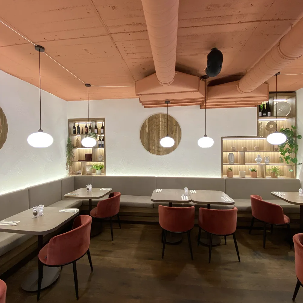 Café-Saal mit einer Tischreihe und einer Sitzbank entlang der Wand, in den Nischen über der Bank stehen Flaschen und Geschirr auf Regalböden, kugelförmige Hängeleuchten