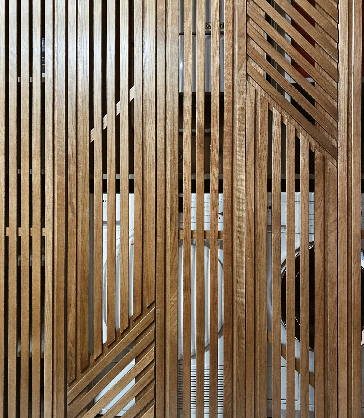 Nahaufnahme einer Front aus vertikalen Eichenlamellen in geometrischem Muster, hinter dem Glas sind Umrisse von Geräten zu erkennen