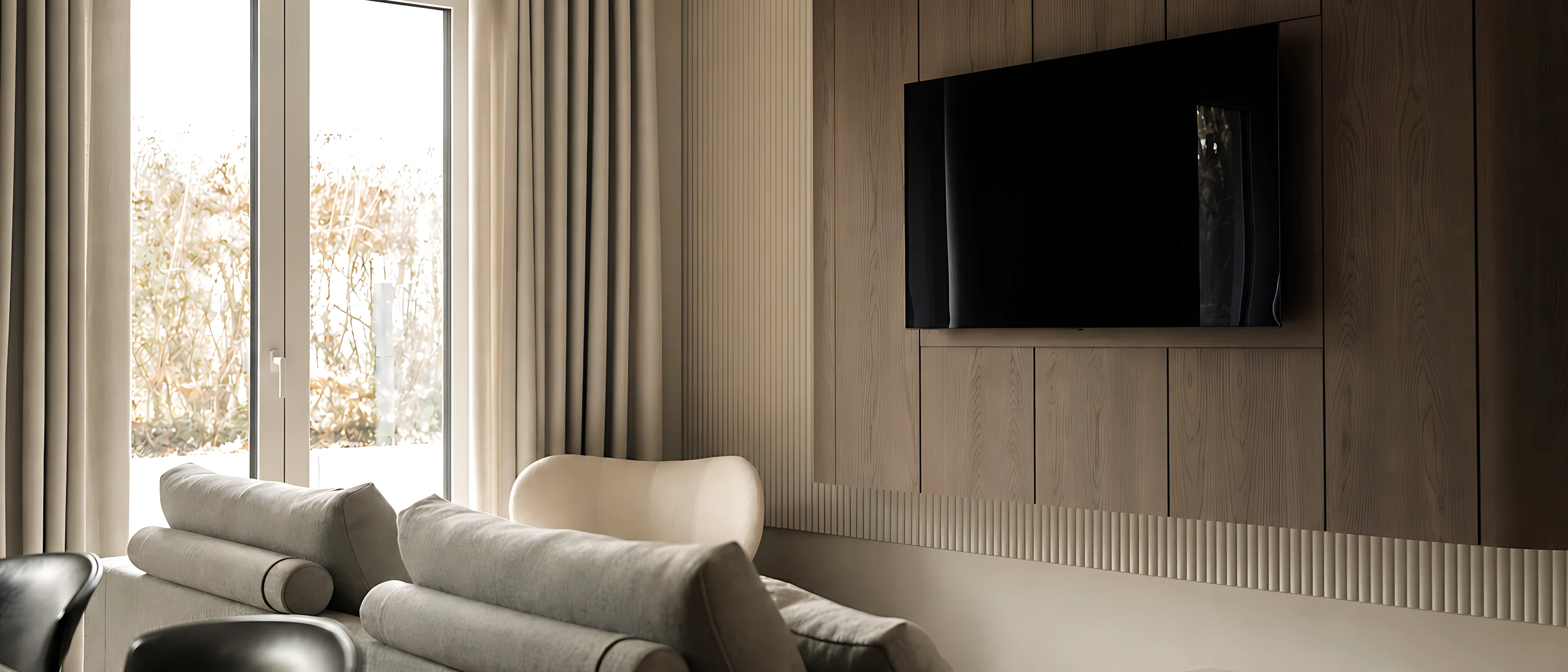 Wohnraum mit holzverkleideter Wand und eingelassenem Fernseher, links ein bodenlanger Vorhang am Fenster, davor helle Sessel